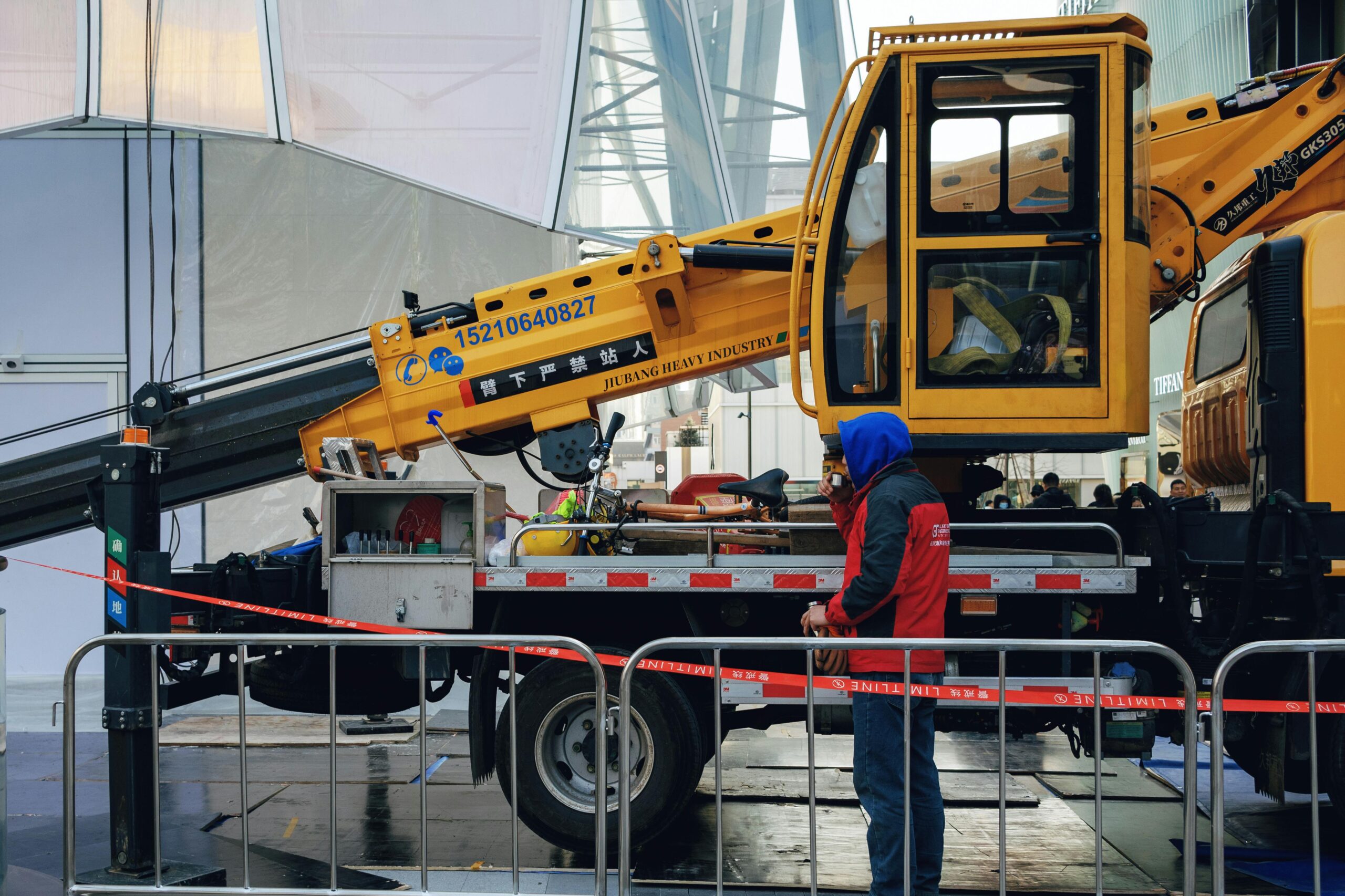 A construction worker wearing a blue hood stands next to a yellow crane truck at a worksite.